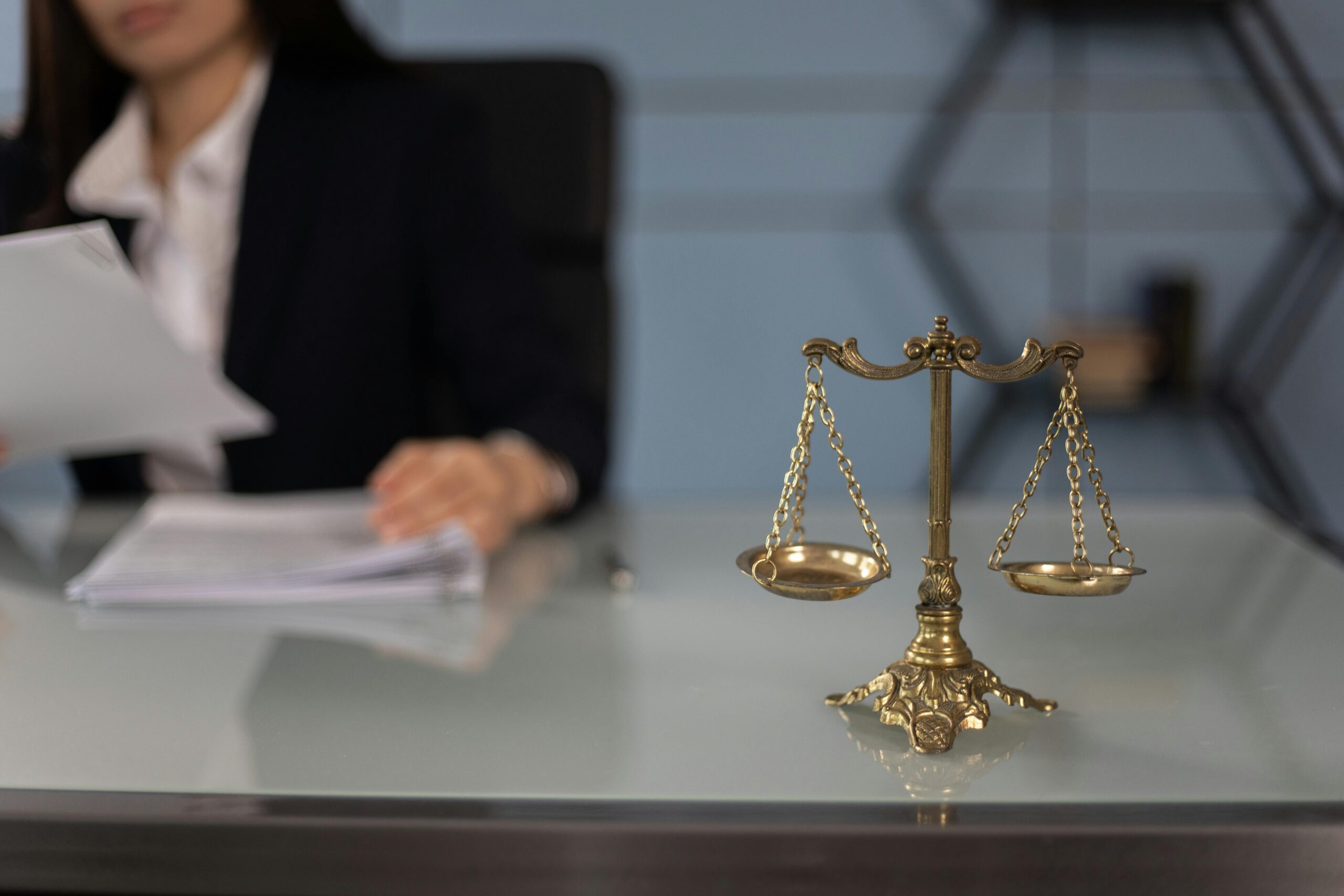 Адвокат по адміністративному праву м.Київ Balance scales on a desk in a professional office with a blurred businesswoman in the background.